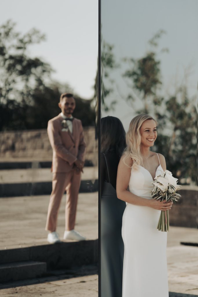 Braut in weißem Kleid mit Blumenstrauß, Bräutigam im rosa Anzug im Hintergrund.