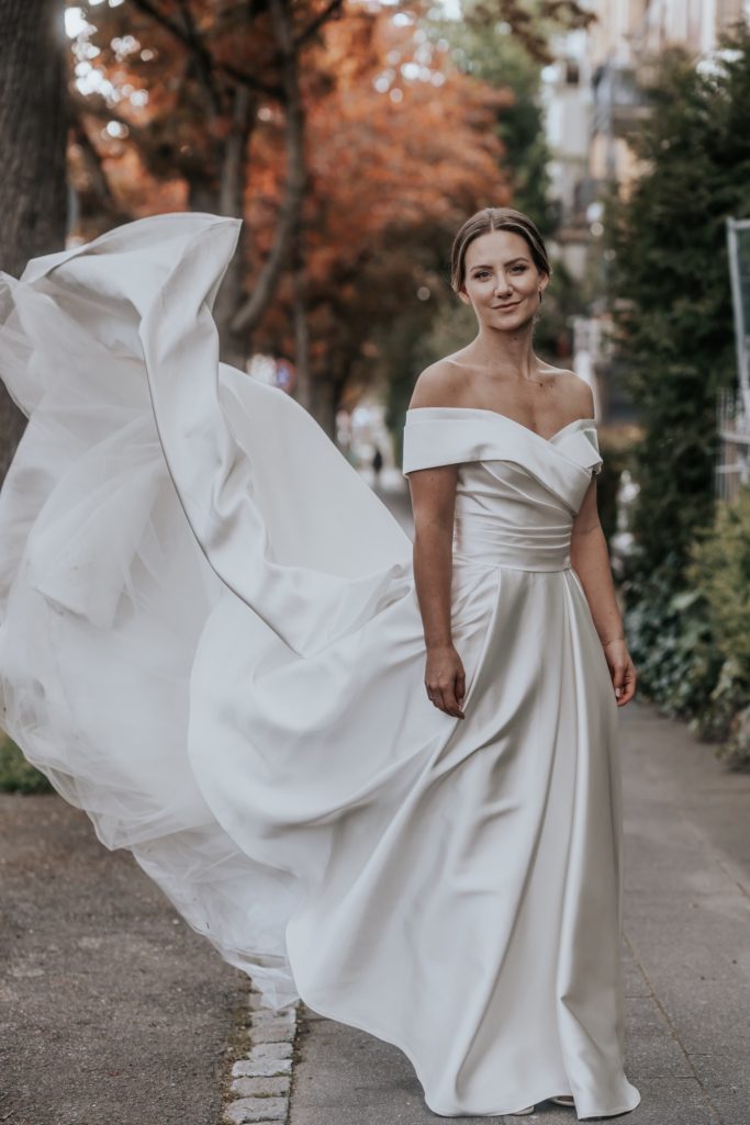 Frau in einem eleganten, schulterfreien Hochzeitskleid auf einem Waldweg.